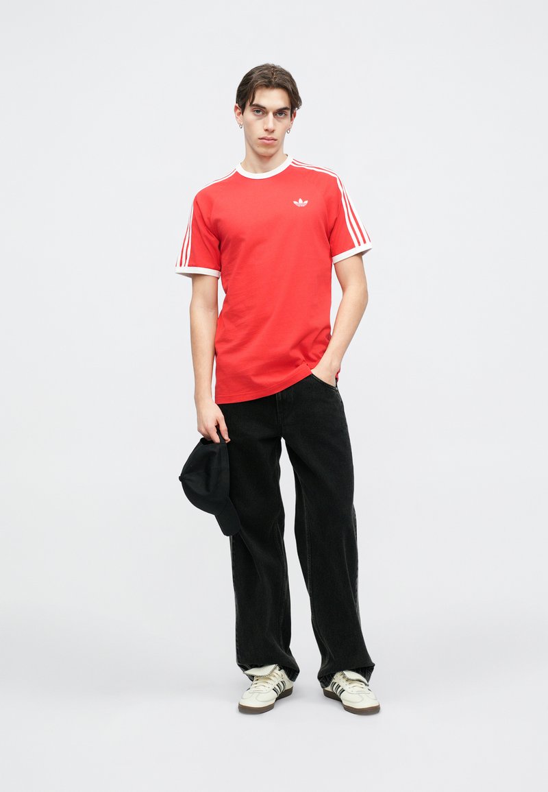 Maglietta rossa con collo a giro bianco e tre strisce bianche sulle maniche, abbinata a jeans neri a gamba larga e sneakers bianche con dettagli neri.