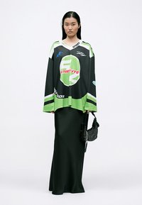 Camisola oversized preta e verde em estilo jersey com logótipos desportivos, combinada com um vestido longo preto. Inclui uma pequena bolsa de malha preta.