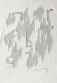 T-shirt en coton de couleur claire, ornée de texte réfléchissant argenté et d'un motif abstrait. Le design comprend des lettres stylisées et des phrases.