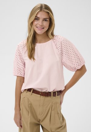Femme souriante aux longs cheveux blonds portant un chemisier rose clair à manches ballon ajourées et un pantalon beige taille haute avec une ceinture marron.