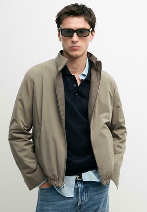 Giovane uomo che indossa una giacca beige, una polo nera, una camicia azzurro chiaro sotto, jeans blu e occhiali da sole neri, in piedi contro uno sfondo semplice.