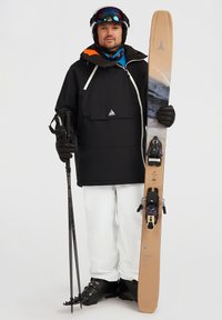 Giacca da sci nera con tasca frontale, pantaloni da sci bianchi, scarponi da sci neri, bastoni da sci e sci beige con attacchi caratterizzati da accenti neri.