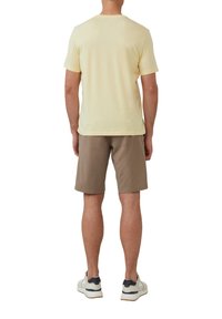 Lichtgele katoenen T-shirt met korte mouwen, gecombineerd met een paarse korte broek. De drager heeft witte en marineblauwe sneakers aan.