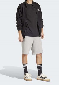 Svart zip-up jacka, svart T-shirt, grå shorts; svarta strumpor med vita ränder; beige sneakers med svarta detaljer och snören.