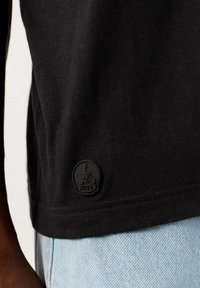 T-shirt nera in cotone con un patch logo JOTT texturizzato sul lato inferiore, abbinata a tessuto denim leggero blu.