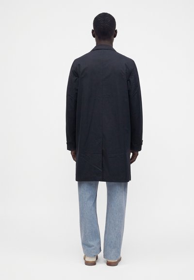 Personne portant un long manteau noir, un jean bleu clair et des chaussures beiges, debout de dos sur un fond blanc uni.