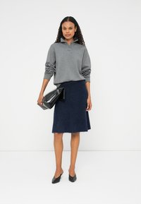 Šedý polo s dlouhým rukávem a límečkem, spárovaný s námořnickou huňatou sukní ke kolenům, držící černou psaníčko a oblečené v černých špičatých botách.