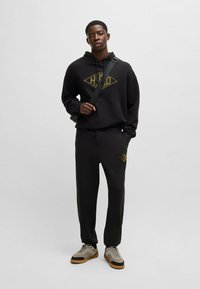 Sort hættetrøje og bukser med guld logo; blødt stof; standard pasform; snørebånd i taljen på bukserne; parret med grå og brune sneakers.