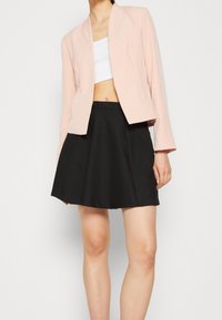Žena v světle růžovém blejzru přes bílý krátký top a černou sukni s vysokým pasem, stojící před jednoduchým bílým pozadím.