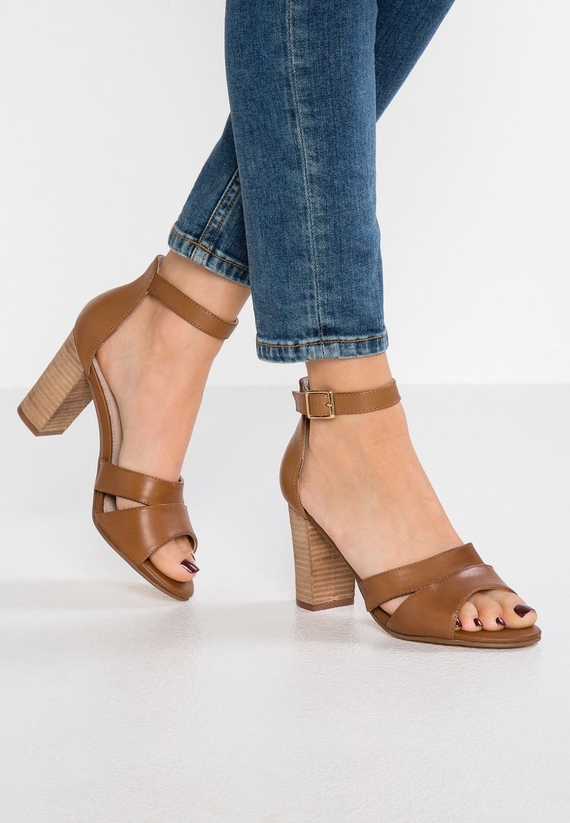 Sandales marron en cuir à talons carrés avec brides à la cheville, portées avec un jean bleu retroussé, sur un sol clair et un fond blanc.