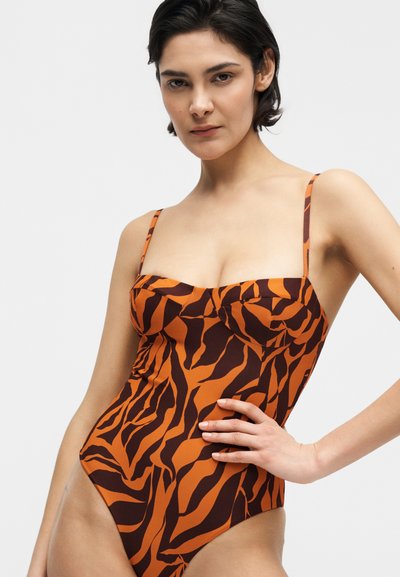 Γυναίκα με κοντά σκούρα μαλλιά, ντυμένη με ολόσωμο μαγιό πορτοκαλί και σκούρου καφέ ριγέ σαν ζέβρα, ποζάρει με το χέρι στη μέση μπροστά σε απλό φόντο.