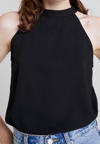 Haut noir sans manches avec un col montant et une texture lisse. Présente une coupe ajustée, un léger drapé, et se porte avec un jean denim bleu clair.