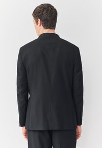 Fekete öltönykabát hátulnézetből, testhezálló szabással, egyoldalas sliccel, sima textúrával és enyhe fényes felülettel.