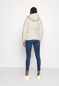 Giacca imbottita color beige chiaro con cappuccio, caratterizzata da sezioni trapuntate. Abbinata a jeans skinny blu scuro e sneaker bianche.