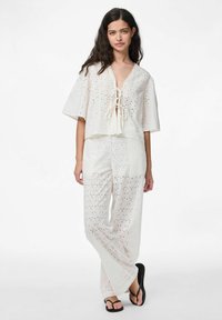 Bílé očkové látkové dvoudílné oblečení s krátkým topem se šněrováním vpředu a širokými kalhotami se stejnými detaily. Černé šlapky.