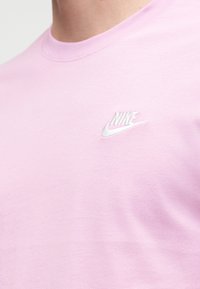 Inimese lähivõte, kes kannab heledat roosat Nike T-särki väikese valge Nike logoga vasakul rinnal.