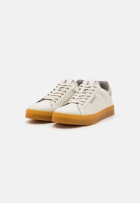 Sneakers bianchi in pelle con finitura testurizzata, stringhe sottili e suola in gomma marrone chiaro. Presentano una fodera in mesh grigia e un logo di marca.