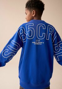 Kungligt blå sweatshirt med stor vit grafisk text på baksidan. Tillverkad av ett mjukt material med ribbade muddar och nederkant.