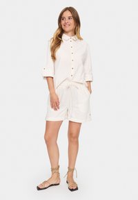 Chemise blanche à boutons avec les manches retroussées, associée à un short crème avec une taille à cordon. Porté avec des sandales à lanières marron.