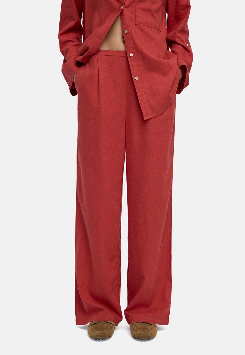 Czerwone spodnie z wysokim stanem i szerokimi nogawkami w połączeniu z dopasowaną koszulą zapinaną na guziki, ręce w kieszeniach, oraz brązowe zamszowe buty na jednolitym tle.