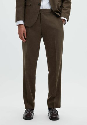 Homme portant un pantalon de costume marron avec un blazer assorti, une chemise blanche, des chaussettes noires et des chaussures habillées en cuir marron foncé, debout sur un fond clair.