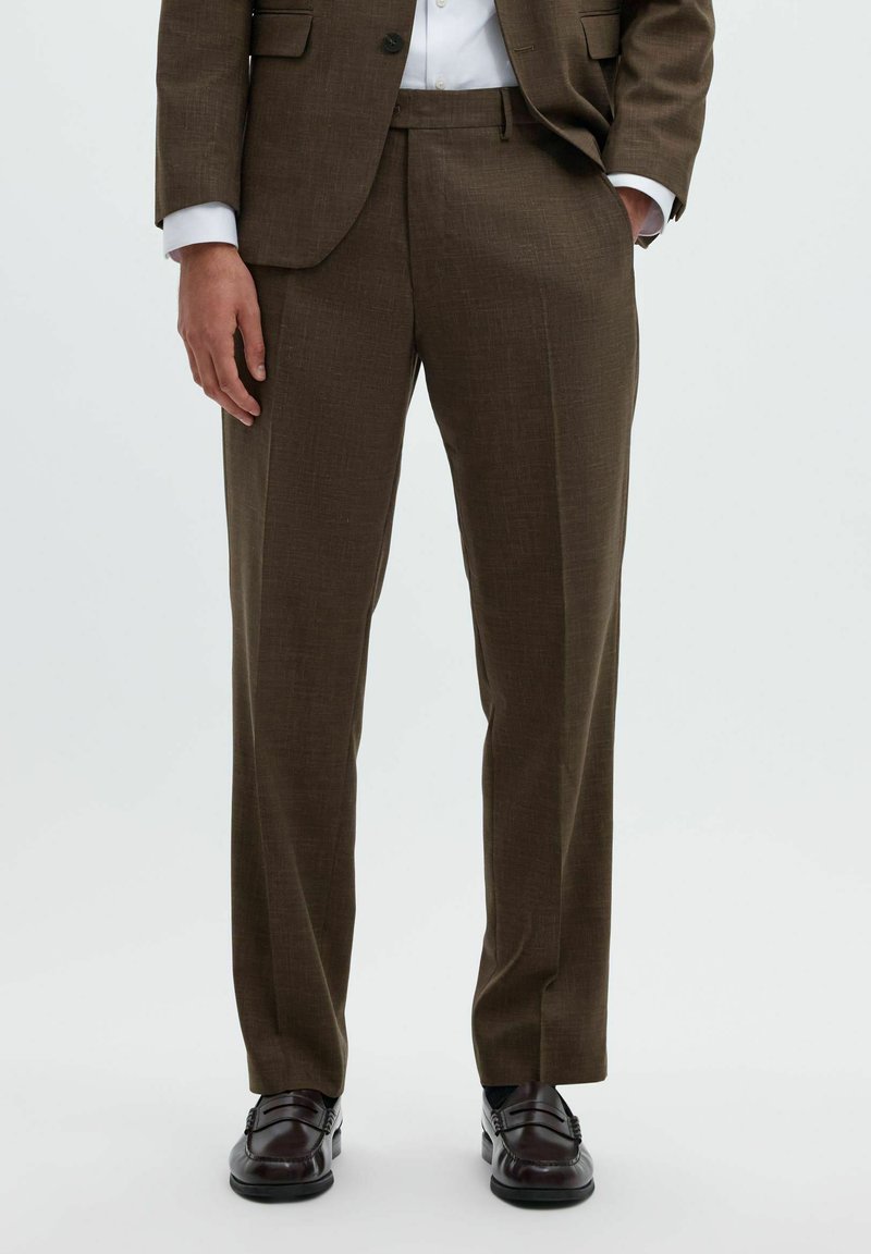 Homme portant un pantalon de costume marron avec un blazer assorti, une chemise blanche, des chaussettes noires et des chaussures habillées en cuir marron foncé, debout sur un fond clair.