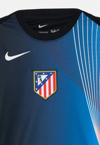Tricou Nike de culoare albastru închis cu guler negru. Prezintă linii diagonale roșii și albe. Pe față se află logo-ul scutului Atletico Madrid.
