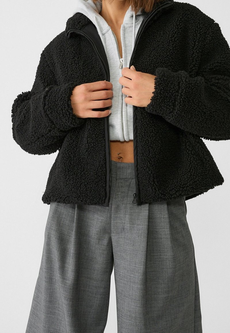 Personne portant une veste noire duveteuse par-dessus un sweat à capuche gris zippé, un pantalon gris taille haute à jambes larges et un piercing au nombril visible.
