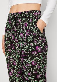 Pantalones florales negros con un patrón morado y verde, cintura elástica, detalle de cordón y bolsillos laterales. Textura de tela suave.