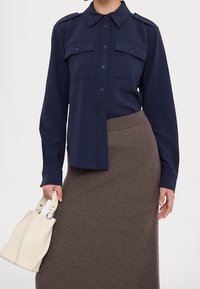Chemise bleu marine à boutons avec deux poches poitrine et épaulettes, assortie à une jupe en tricot côtelé marron et à un petit sac à main blanc.