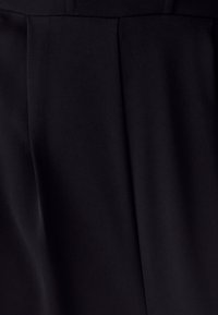 Artimas juodo audinio vaizdas su vertikaliais raukiniais ir subtilia įstrižine tekstūra švelnioje šviesoje.