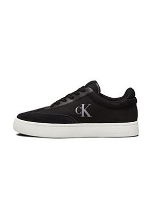 Zapatilla de ante negra con suela blanca, con ojales y un logo "CK" gris en el costado, diseñada con una textura suave y forma redondeada.