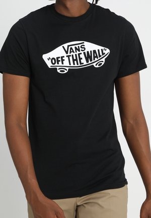Person trägt ein schwarzes Vans T-Shirt mit weißem Skateboard-Logo, auf dem "Vans Off The Wall" steht, und beige Hosen.