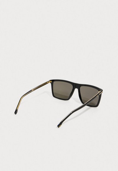 Lunettes de soleil rectangulaires noires avec des verres foncés et des branches rehaussées d'or, posées ouvertes sur un fond clair.