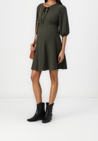 Vestido verde oscuro, hasta la rodilla, con mangas abullonadas de 3/4 y detalle de lazo en el cuello. Combinado con botas de cuero negras hasta el tobillo y un bolso cruzado marrón.