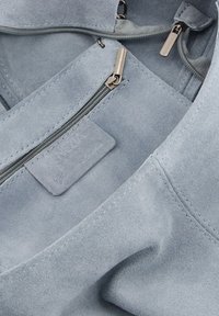 Helleblaue Wildledertasche mit glatter Textur, ausgestattet mit zwei Reißverschlussfächern und einem Etikett mit geprägt Branding.