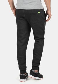 Black cotton sweatpants with a tapered fit, featuring a back pocket and a small green logo accent. Styled with black athletic shoes.