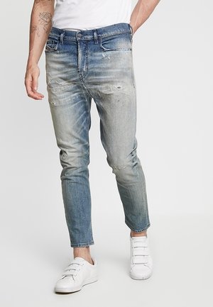Homme portant un jean bleu délavé et usé avec des baskets blanches, debout avec une main dans la poche sur un fond uni.