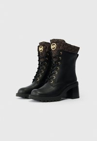 Botas de couro pretas com um salto robusto, apresentando detalhes em ouro, uma bainha de tecido castanho com um padrão de logótipos e atacadores na parte da frente.