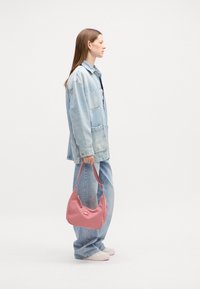 Sac à épaule rose clair avec une texture lisse, doté d'une fermeture éclair supérieure et d'une bandoulière réglable, tenu par une personne en veste en denim et jeans.