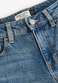 Pantalones cortos de mezclilla de media altura en azul claro, con una tela texturizada, costuras naranjas, botón frontal con una etiqueta que muestra la talla y el origen.
