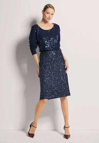 Marineblauwe paillettenjurk met een ronde halslijn en lange mouwen. Aansluitende rok tot aan de knie, versierd met glanzende pailletten. Hakken.