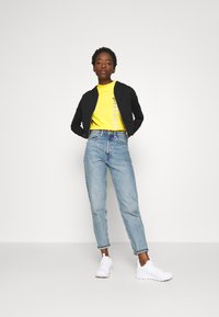 Sudadera negra con cremallera, camiseta amarilla con un gráfico gris, jeans de tiro alto de color azul claro y zapatillas blancas. Diseño sencillo y ajuste casual.