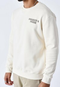 Sweatshirt couleur crème avec des poignets et un ourlet côtelés, présentant le texte en relief "PROJECT X PARIS" en gris sur la poitrine gauche. Texture lisse.