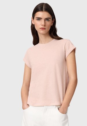 Kobieta o długich ciemnych włosach, ubrana w jasnoróżową koszulkę z krótkim rękawem i białe spodnie, stojąca z rękami w kieszeniach na jednolitym tle.