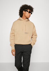 Felpa beige realizzata in tessuto morbido con tasca a marsupio e cappuccio con cordino. Presenta il logo "Calvin Klein Jeans" in nero. Abbinata a pantaloni neri.