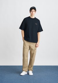 Tmavě modré oversized tričko s malou vyšitou nášivkou, spárované s khaki cargo kalhotami s kapsami a bílými teniskami. Černá čepice na hlavě.