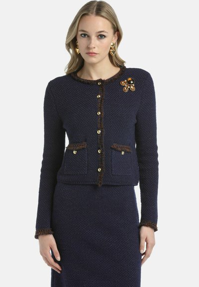 Navy knit jacket with a textured finish, featuring gold buttons, two front pockets, and a decorative flower brooch with brown accents.