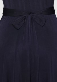 Tissu bleu marine foncé avec un nœud noué en ruban large assorti à la taille, créant de doux plis verticaux.