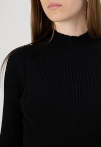Fekete bordázott kötött pulóver magas nyakkal és V-alakú díszítéssel a mellrészen, hosszú ujjakkal és sima textúrával.
